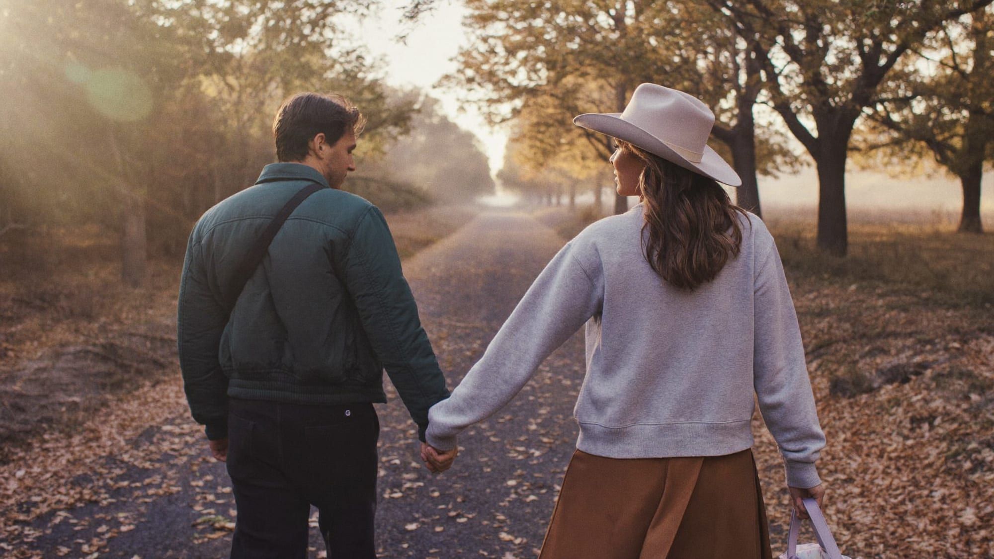 A couple walking hand in hand along a tree-lined autumn path, seen from behind